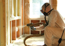 Technician in full hazmat suit with gas mask, applying spray foam insulation in an unfinished wall.